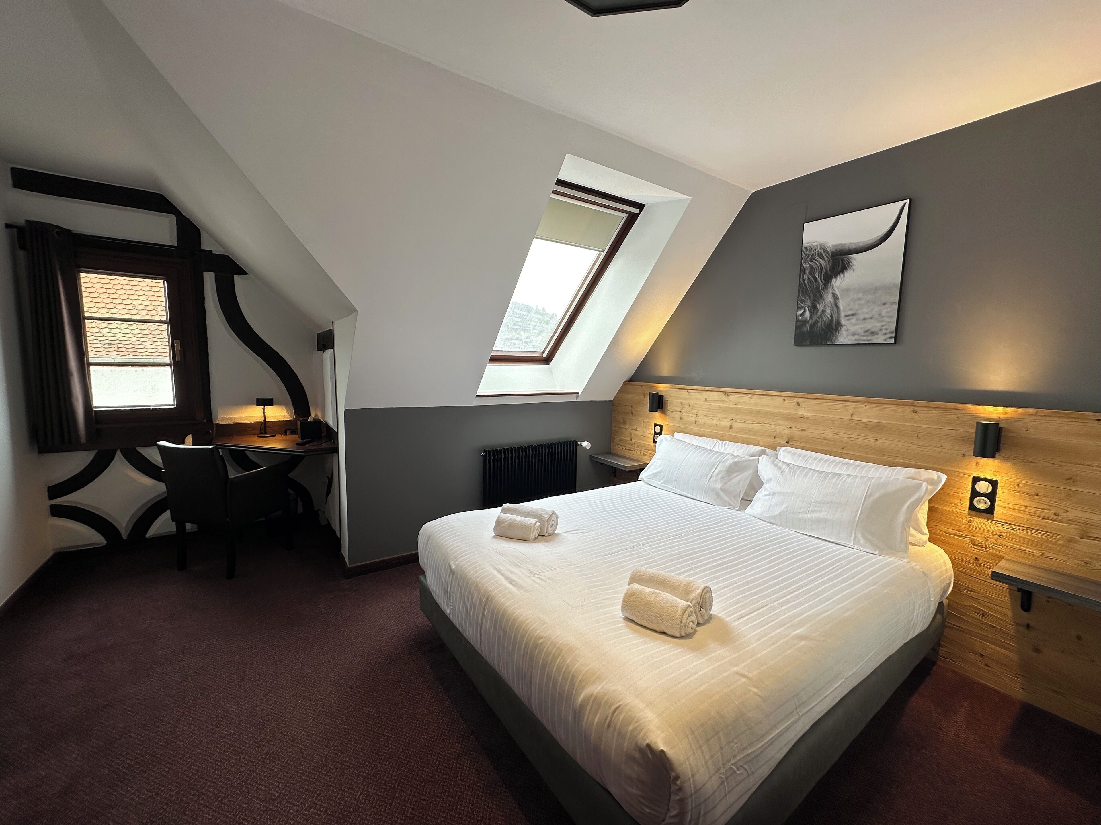 Ein modernes Hotelzimmer mit einem großen Bett, einem Schreibtisch und einem Stuhl. Über dem Schreibtisch befindet sich ein Oberlicht und an der Wand hängt ein Gemälde.