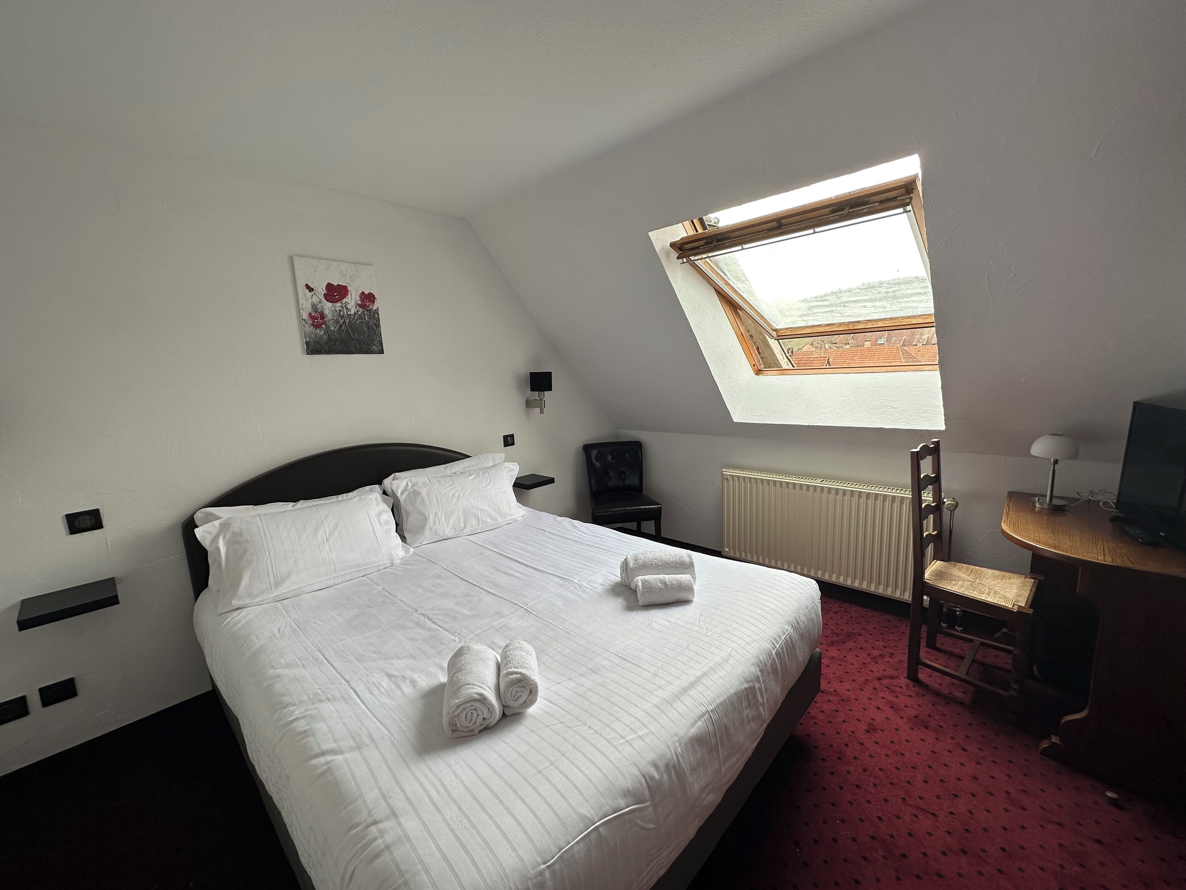 A neatly made bed with white linens and two rolled towels, a small desk with a lamp and a television, a radiator, and a skylight above the bed.
