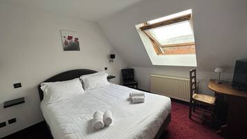 A neatly made bed with white linens and two rolled towels, a small desk with a lamp and a television, a radiator, and a skylight above the bed.
