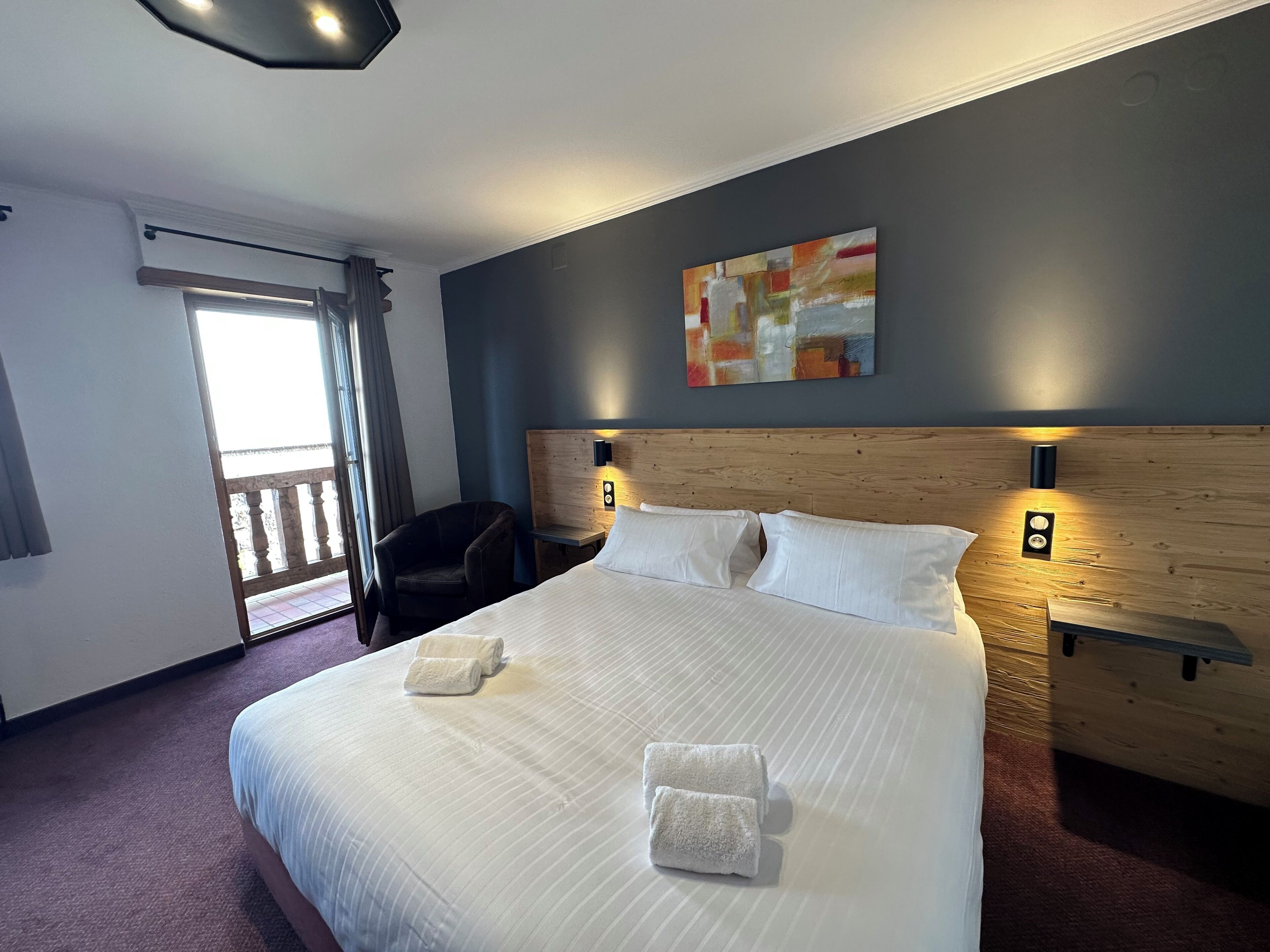 Ein Hotelzimmer mit einem großen Bett, zwei Nachttischen, einem Sessel, einem Bild an der Wand und einer Balkontür.