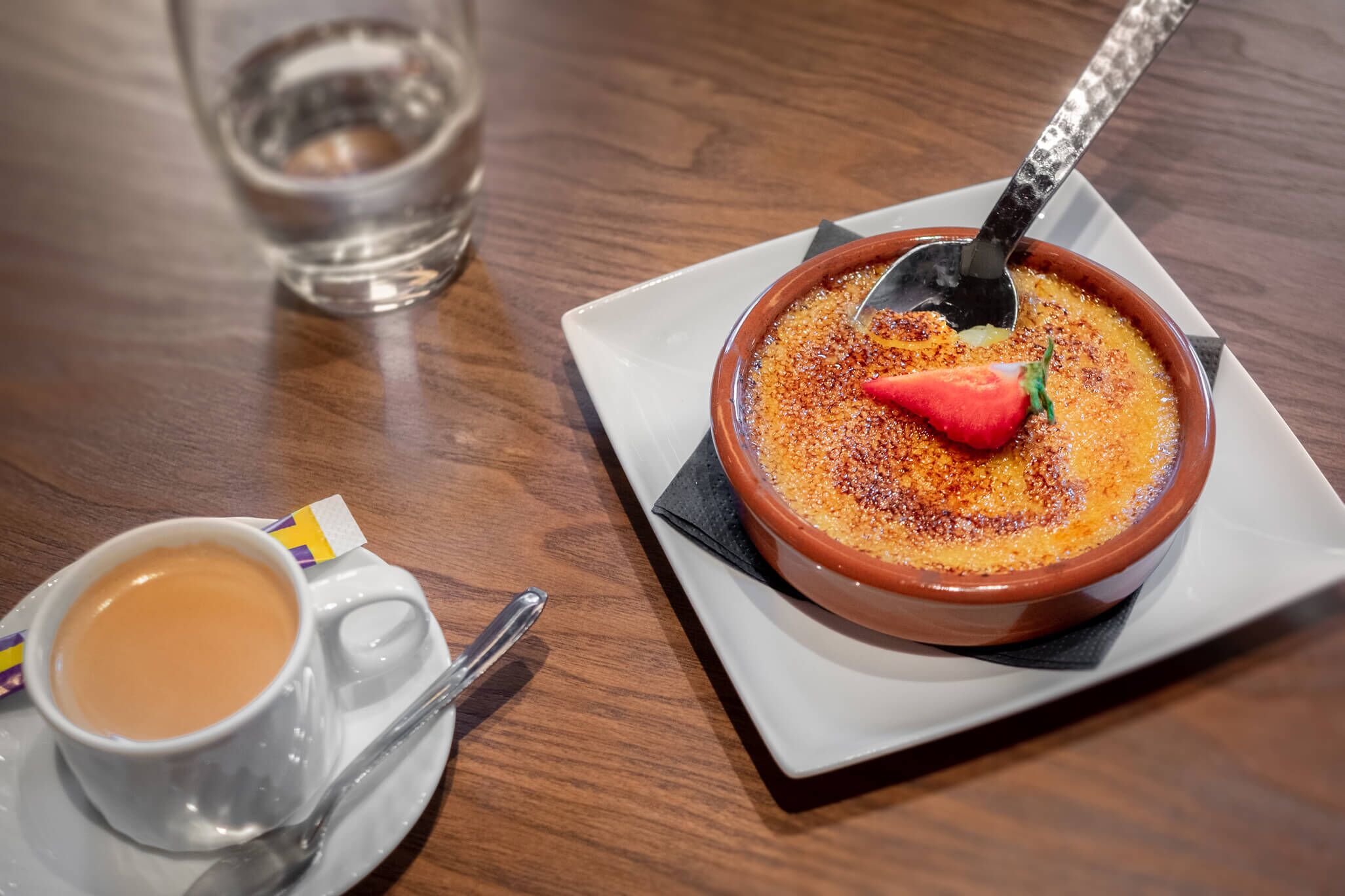 Un plato de postre con una fresa encima, una taza de café y un vaso de agua sobre una mesa de madera.