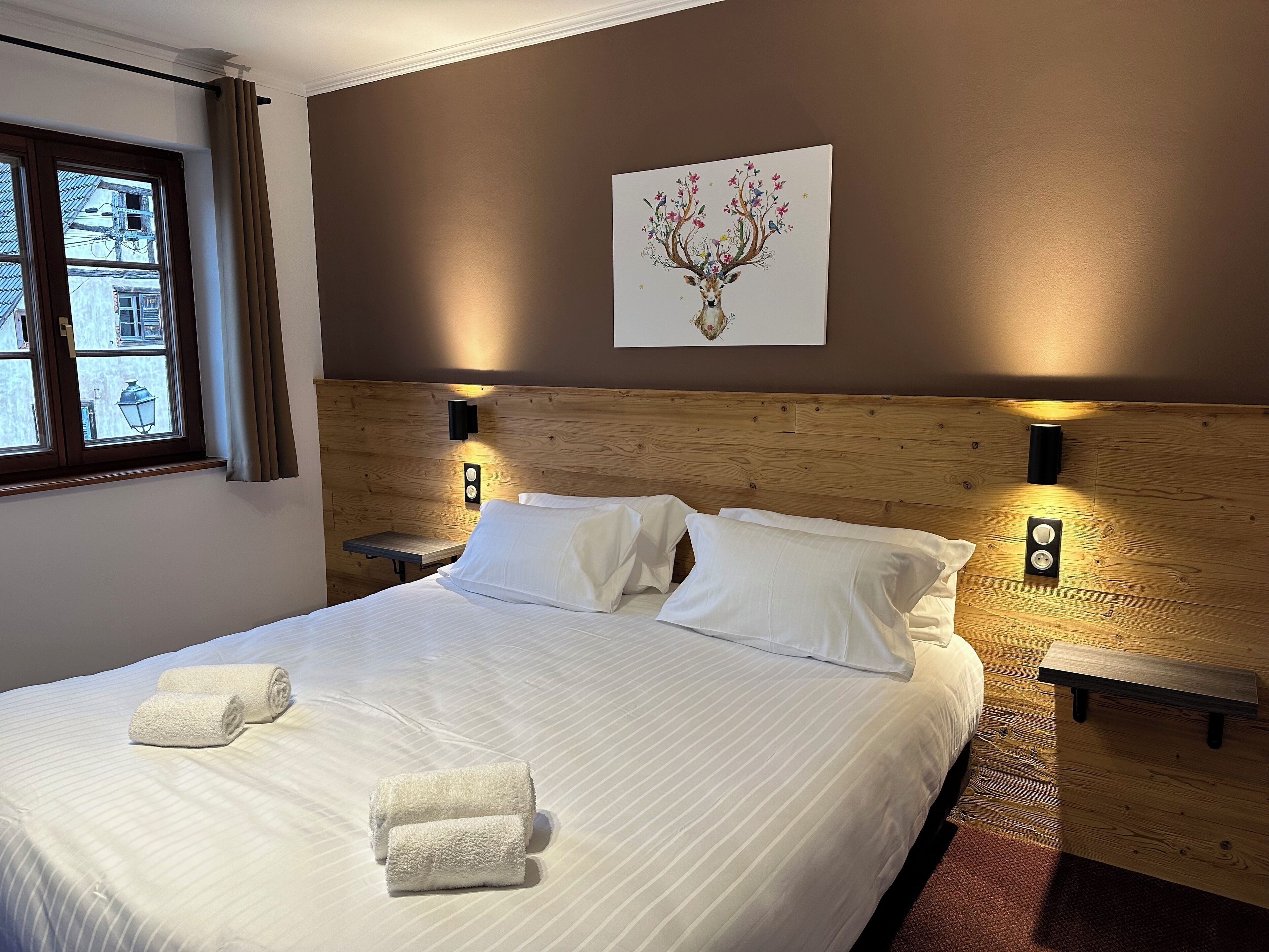 A neatly made bed with white linens and rolled towels, a wooden headboard, two bedside tables with wall-mounted lamps, and a window with curtains.
