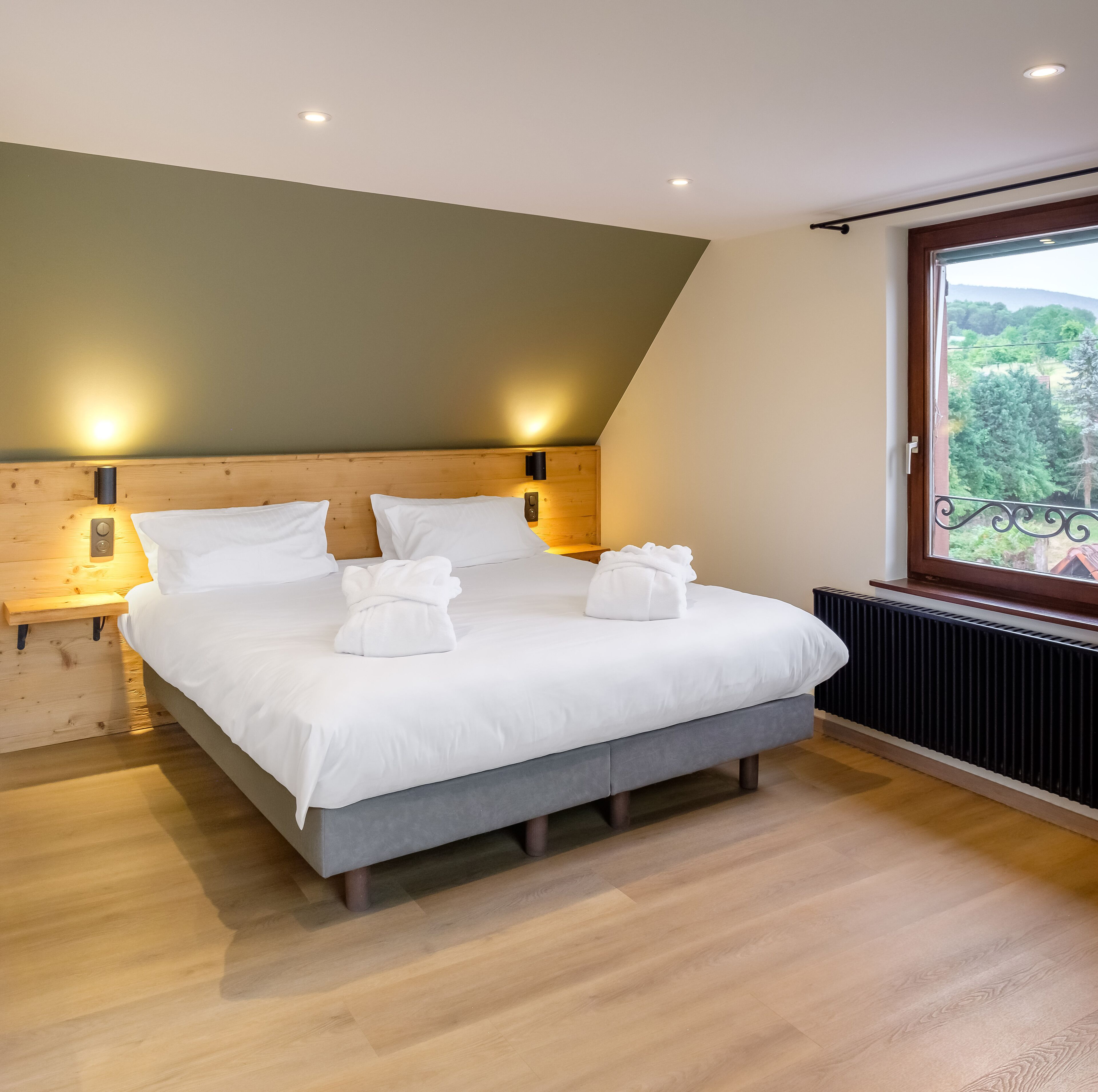 Un dormitorio moderno con cabecera de madera, una cama grande con ropa de cama blanca, dos lámparas de noche y vistas a zonas verdes a través de una ventana.