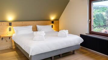 Un dormitorio moderno con cabecera de madera, una cama grande con ropa de cama blanca, dos lámparas de noche y vistas a zonas verdes a través de una ventana.