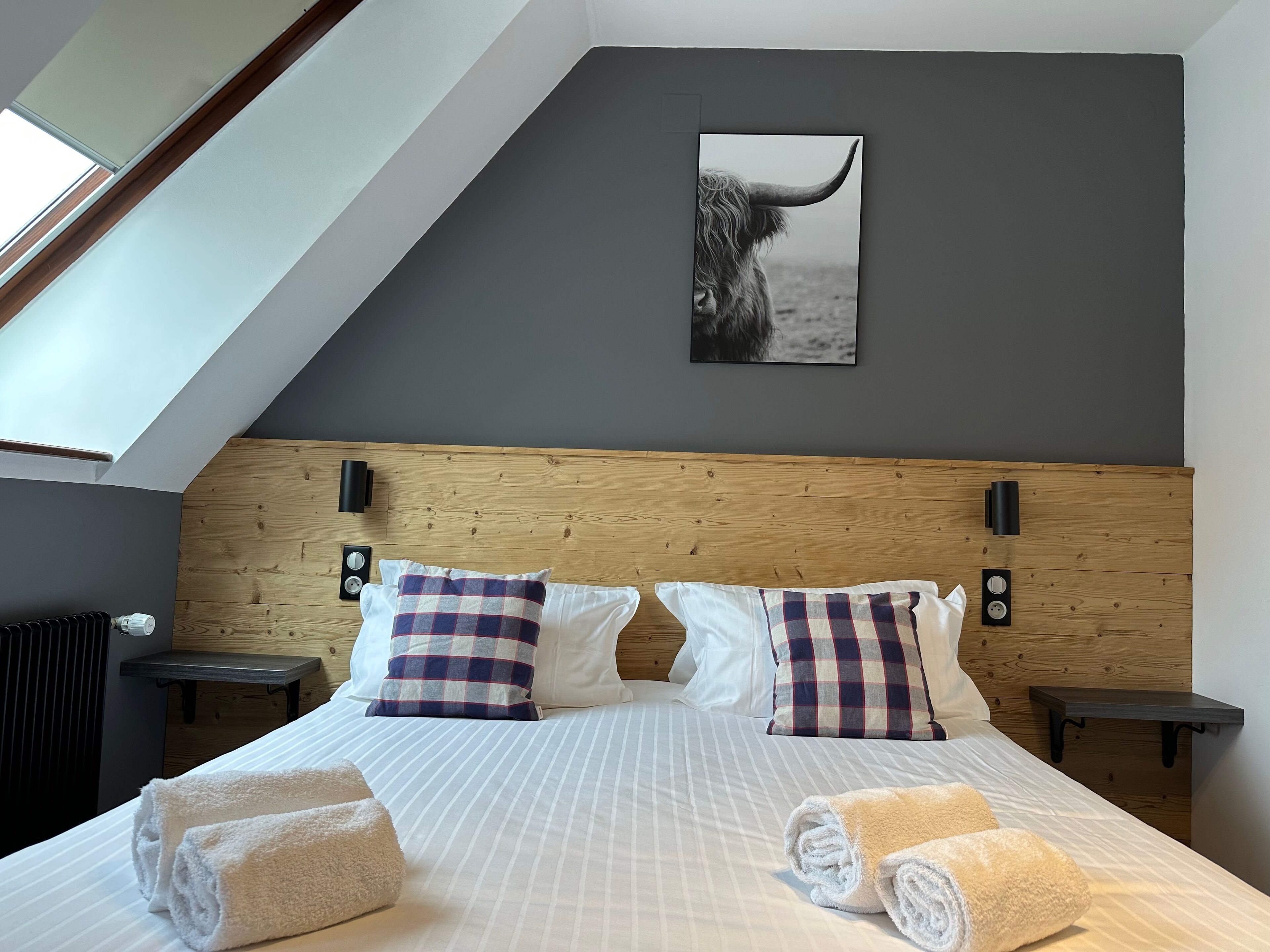 Ein modernes Hotelzimmer mit schräg versiegelter Decke, einem großen Bett mit gestreifter Bettwäsche, zwei karierten Kissen und einem aufgehängten Bild eines Stiers.
