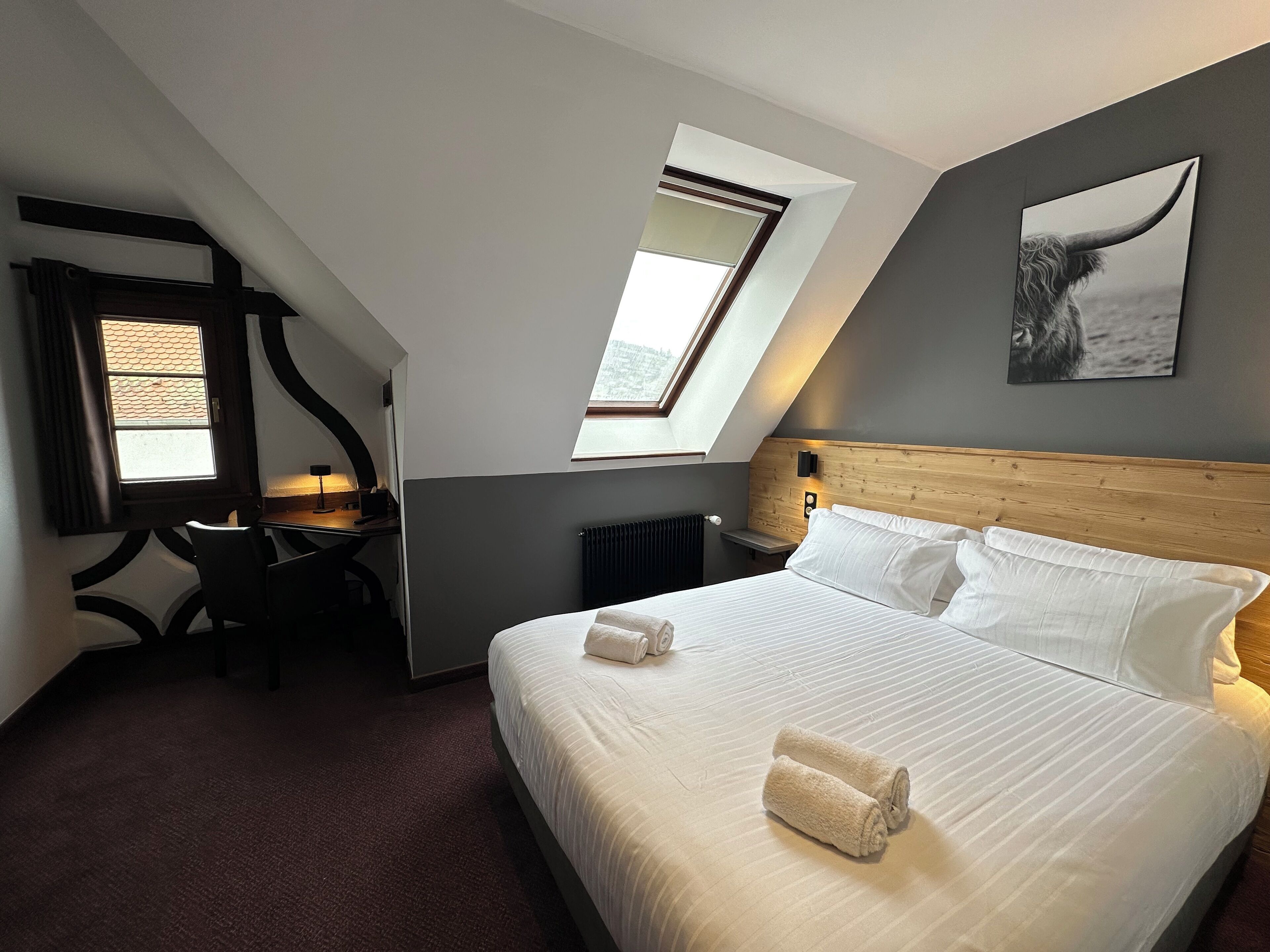 A modern hotel room with a large bed, two bedside tables, a desk, and a chair. There are two windows providing natural light, and a painting of a whale on the wall.