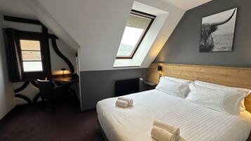 A modern hotel room with a large bed, two bedside tables, a desk, and a chair. There are two windows providing natural light, and a painting of a whale on the wall.