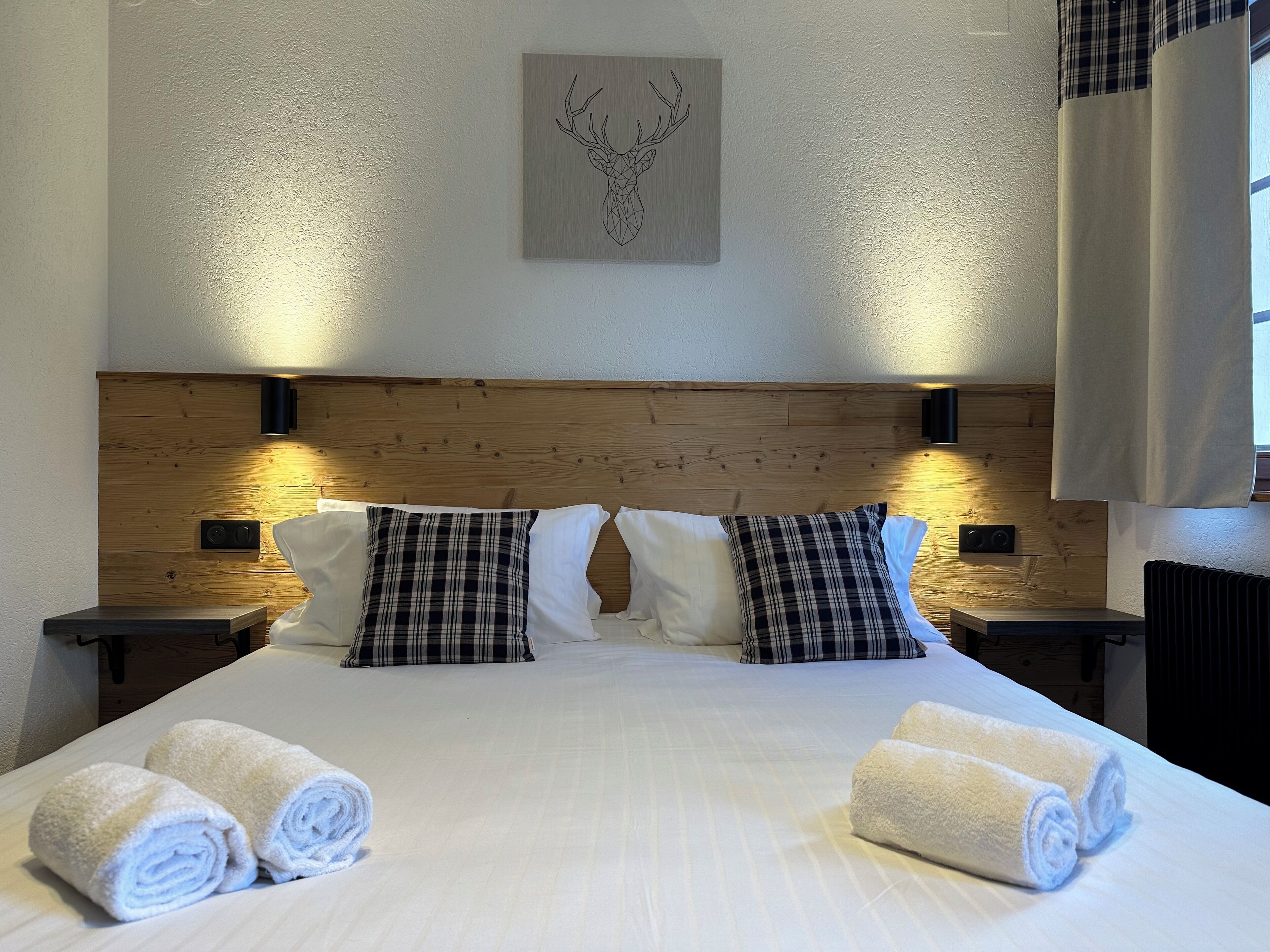 Een opgemaakt bed met geruite en effen kussens, twee nachtkastjes met lampen, en een wanddecoratie van een hertenkop.