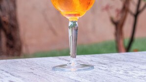 A glass of orange beverage with a straw, placed on a table with a blurred background of greenery and a tree. - Hôtel Au Heimbach (Lembach)