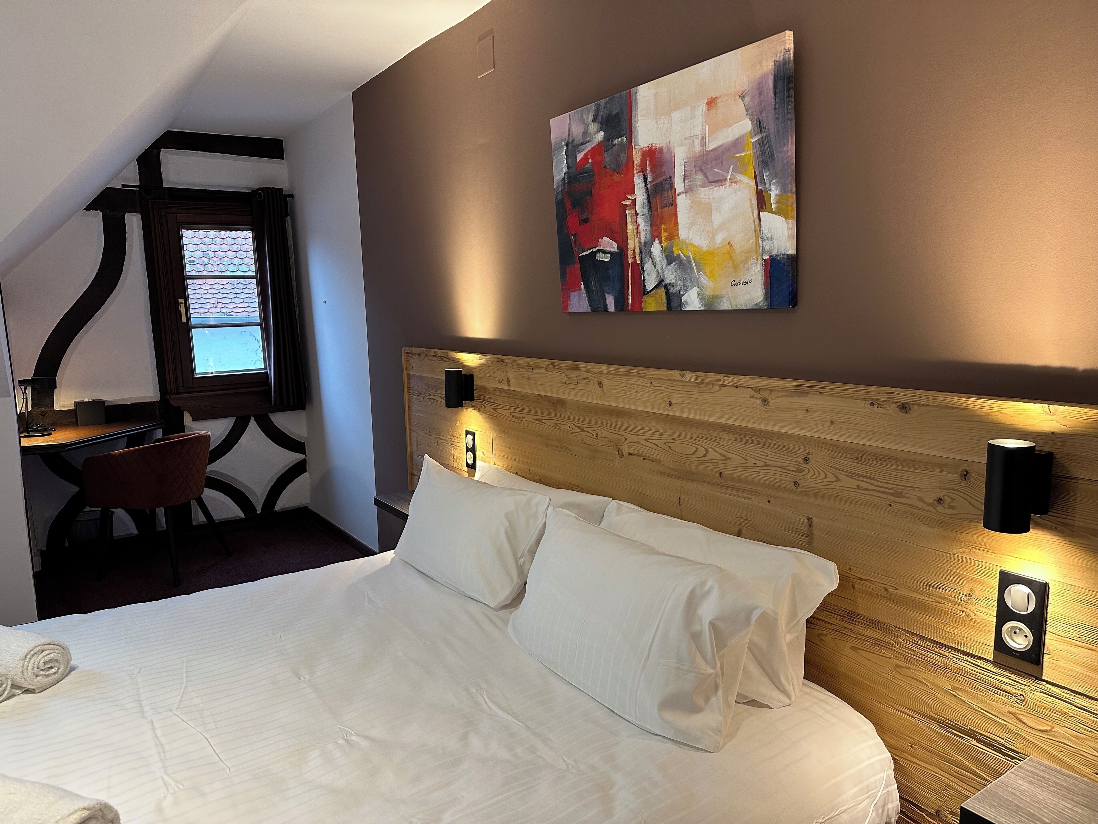 Ein Hotelzimmer mit Bett, Schreibtisch und Stuhl. An der Wand hängt ein Gemälde, und es gibt ein Fenster mit Vorhängen.