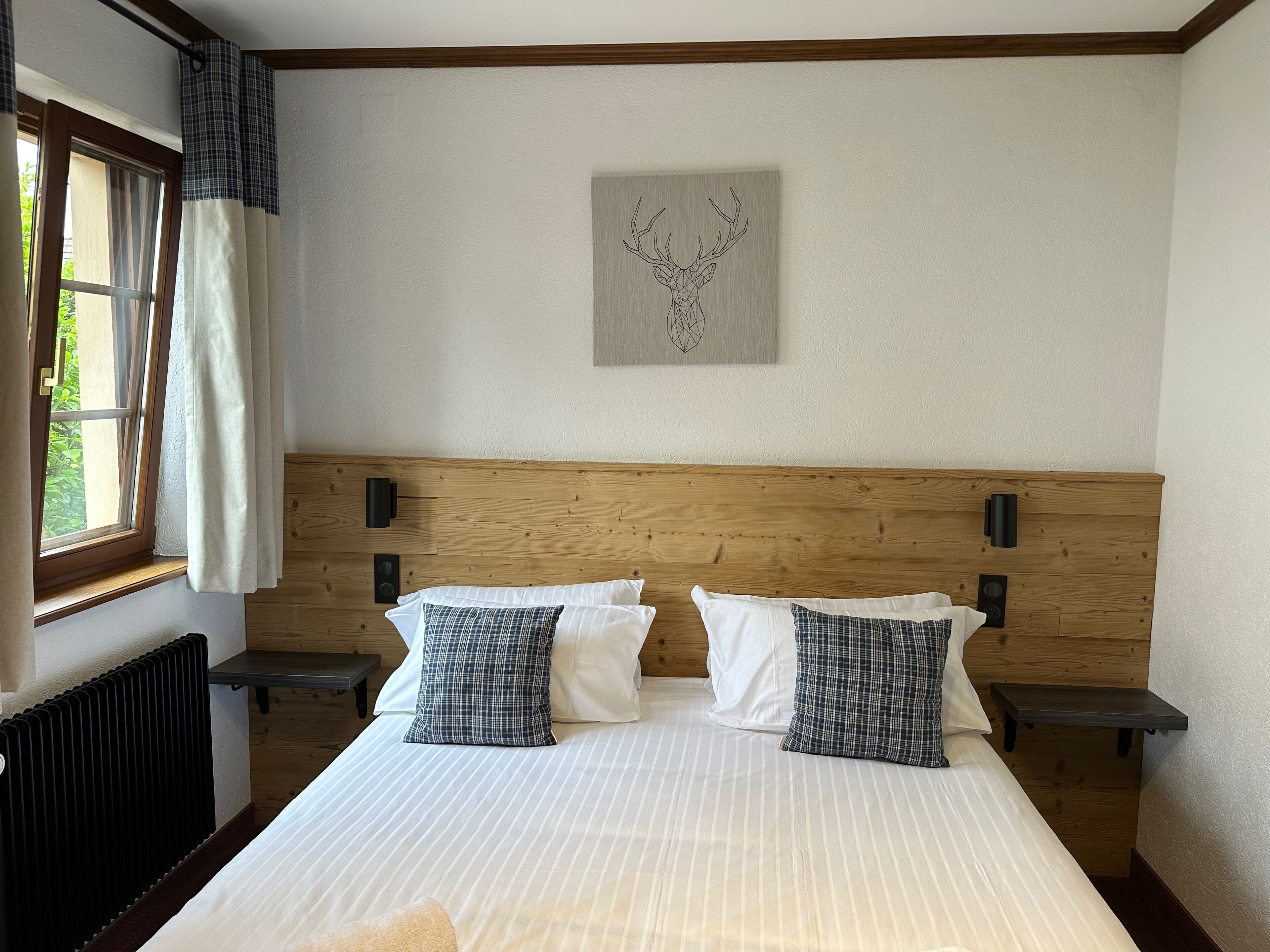 A bedroom with a wooden headboard, two bedside tables, a radiator, and a window with curtains.