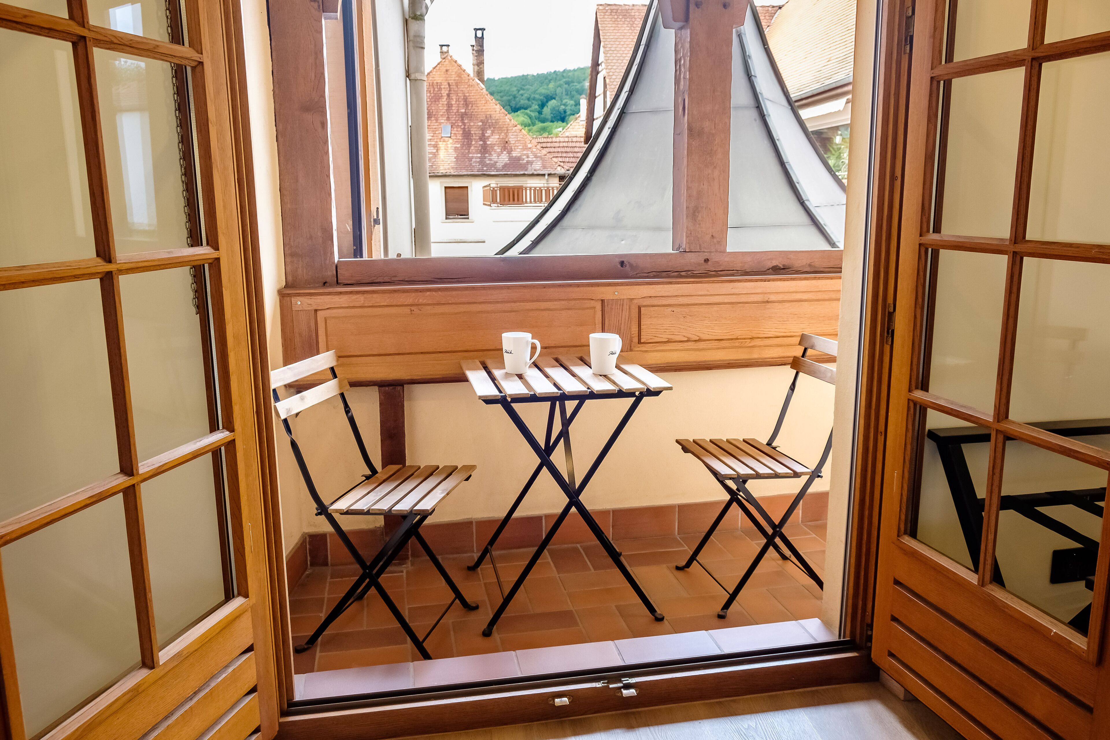 A small balcony with a wooden table and two chairs, a window with wooden frames, and a view of buildings and greenery.