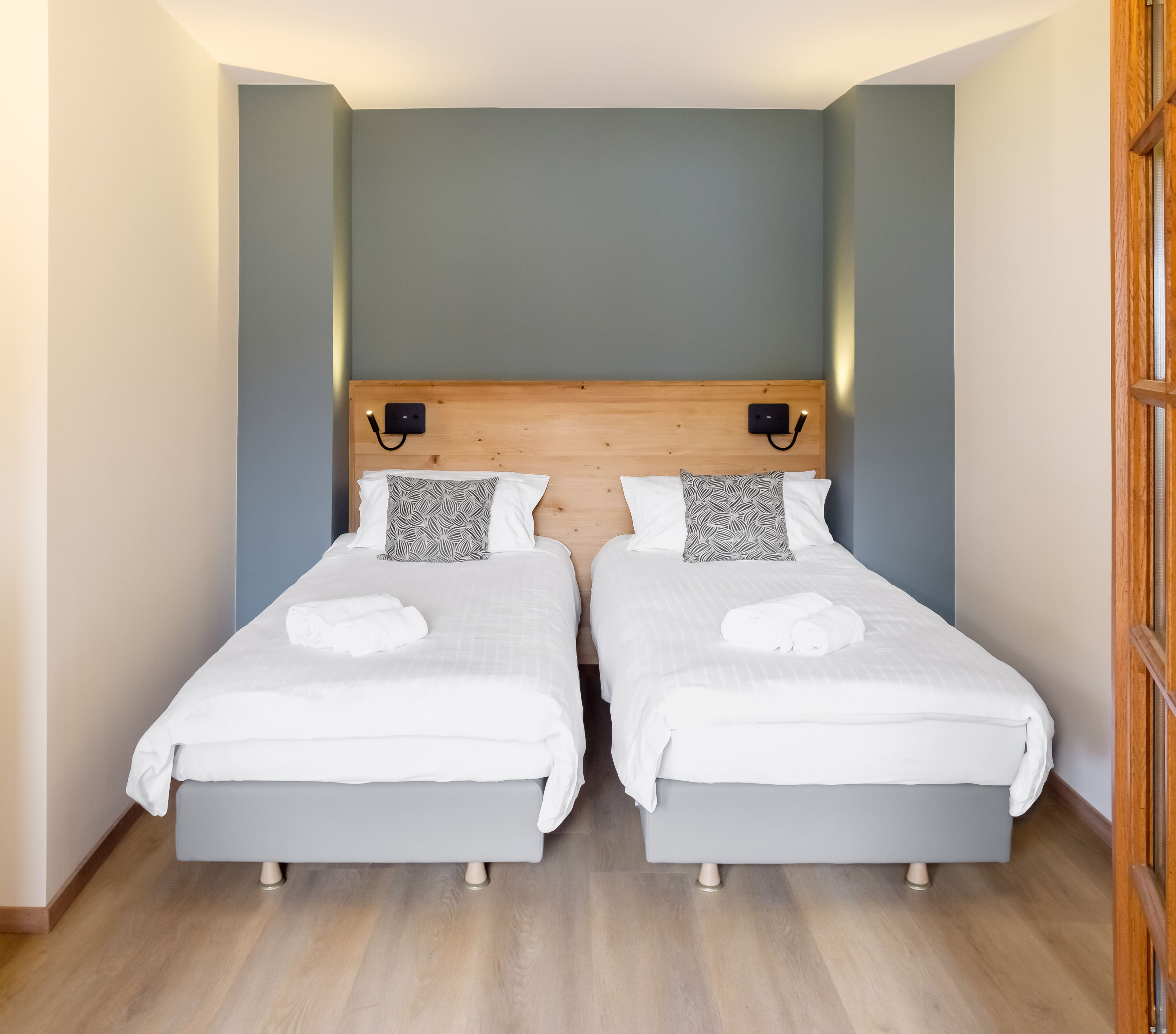 Ein Doppelzimmer mit zwei Betten, jeweils mit weißer Bettwäsche und gemusterten Kissen, einem Holzkopfende und einer kleinen Wandleuchte.