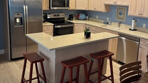A modern kitchen with wooden cabinets, stainless steel appliances, a central island with stools, pendant lighting, and a dishwasher.