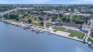An aerial view of a waterfront area with a marina, buildings, and a large open space with a patterned lawn.