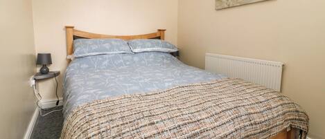 A bedroom with a wooden bed, a patterned blue bedspread, a woven throw blanket, a bedside lamp, and a radiator.