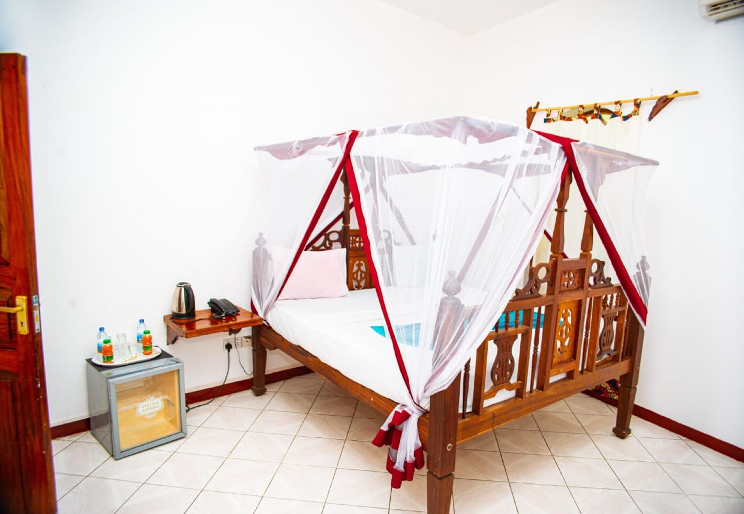 A wooden bed with a mosquito net, a small table with a kettle and bottles, and a refrigerator.