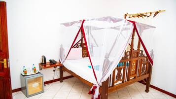 A wooden bed with a mosquito net, a small table with a kettle and bottles, and a refrigerator.