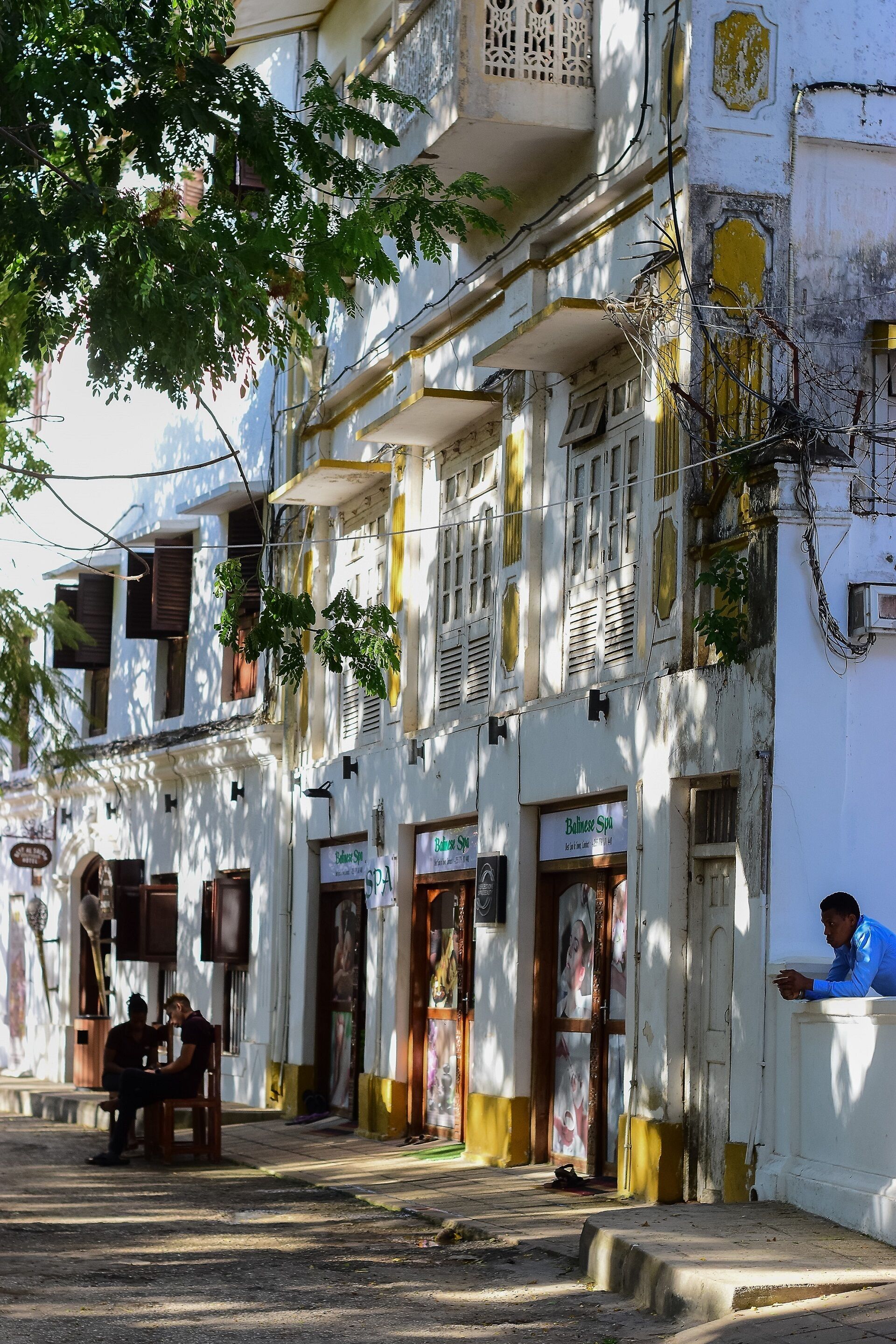 A street view with a building featuring multiple balconies, a shop with a sign, and hanging plants.