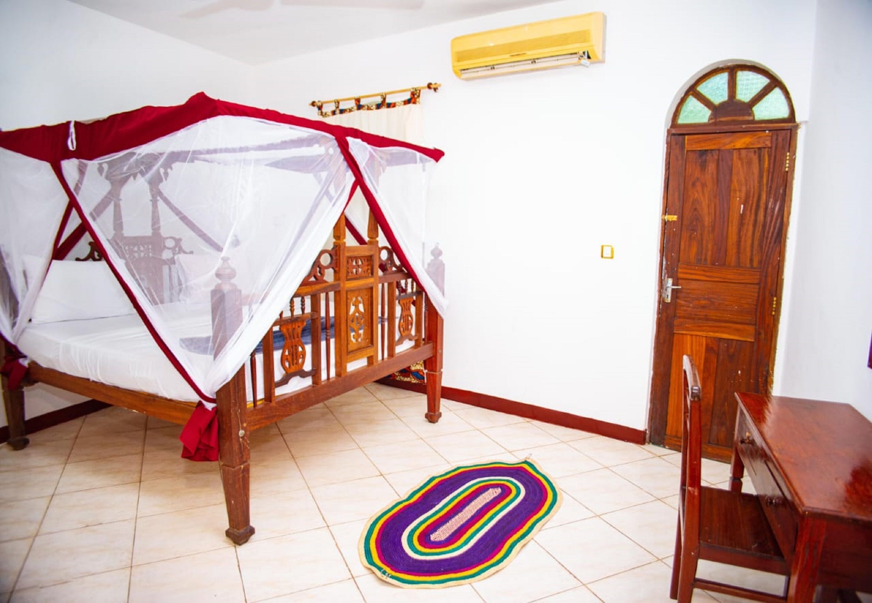 A bedroom with a wooden bed, a mosquito net, a wooden chair, a wooden desk, and a door.