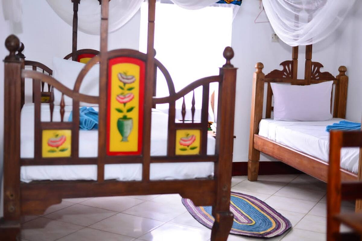 Een kamer met twee houten bedden, waarvan één met hemelbed en één zonder. Op de vloer ligt een kleurrijk tapijt.