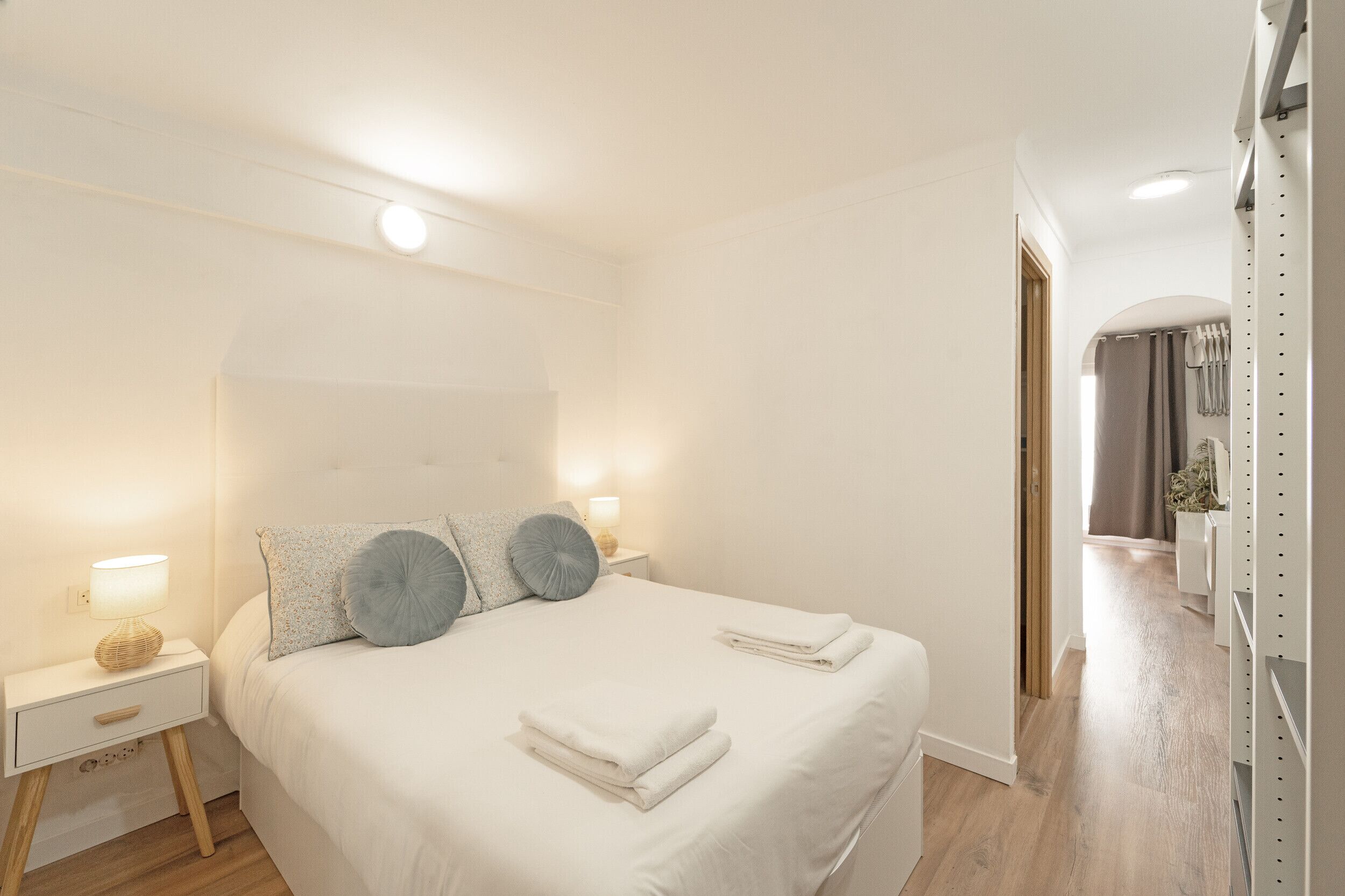 Een net opgemaakt bed met kussens, een bedlampje en een opgevouwen handdoek op een witte nachtkastje.