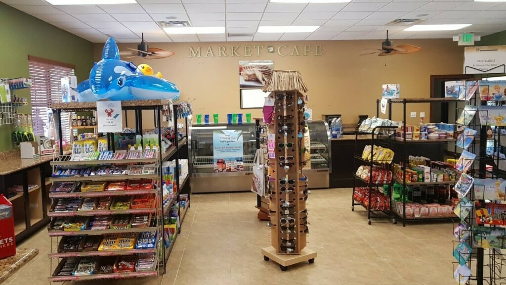 Un café situé sur un marché, avec des étagères remplies d’en-cas, un présentoir de lunettes de soleil et un panneau indiquant la vente de cartes postales.
