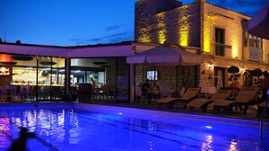 An outdoor swimming pool with lounge chairs and umbrellas, a bar area with a 'Red Bull' sign, and a stone building with a balcony.