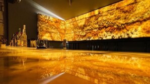 A spacious lobby with a polished floor reflecting the warm lighting. A long reception desk is visible with various items on it.