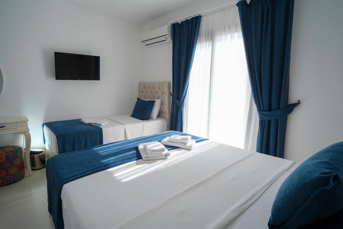 Quarto de hotel com cama, mesa de cabeceira, cadeira, televisão e janela com cortinas.
