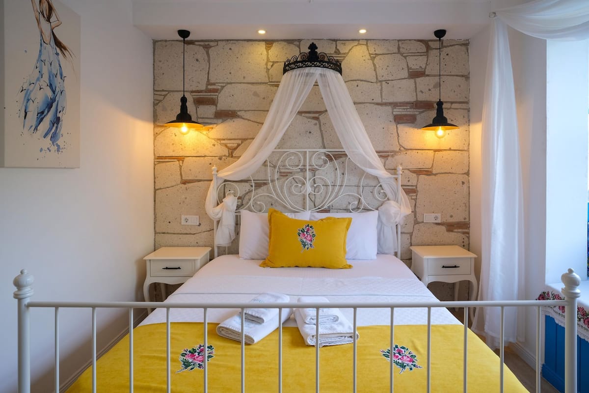 Uma cama com dossel, roupa de cama branca e almofada amarela. Há duas mesas de cabeceira com luminárias.