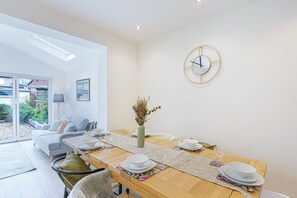 A dining area with a wooden table set for four, a vase with dried plants, and a wall clock.