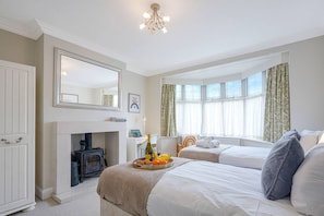 Quarto de hotel com lareira, cama, mesa de cabeceira com garrafa e frutas, e uma janela grande com cortinas.