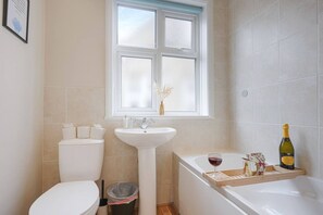 A bathroom with a toilet, sink, and bathtub. There is a window above the sink, and a bottle of champagne and a glass on the bathtub. - Elegant House • Central Location • Sleeps 8 • Free Parking • Fast Wifi • VirginTV • By Yoko Property (Northampton)