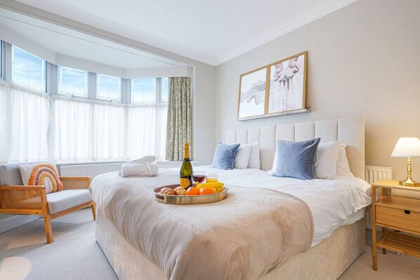 A hotel room with a large bed, a bedside table with a lamp, a chair, a window with curtains, and a tray of breakfast items.