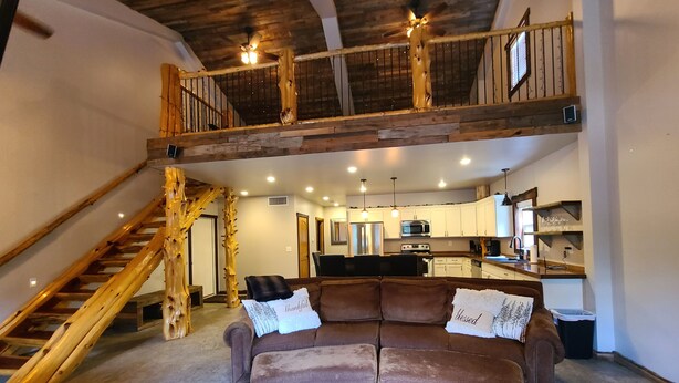 A spacious living area with a high ceiling, wooden staircase, and a kitchen in the background.