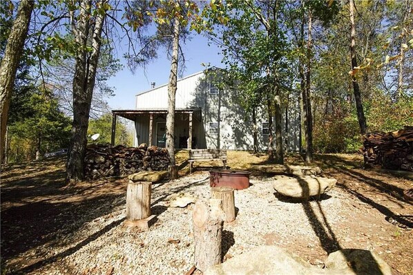 Una cabaña rústica con terraza de madera y chimenea de piedra, rodeada de árboles.
