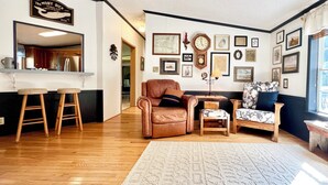 Ein gemütliches Wohnzimmer mit einer braunen Ledersofa, zwei Holzschemeln, einem gemusterten Sessel und einem Holztisch. Eine große Uhr und verschiedene gerahmte Bilder schmücken die Wände.