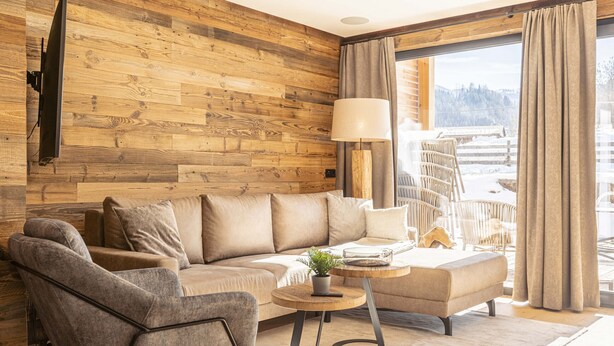 Ein modernes Wohnzimmer mit einer Holzvertäfelung, einer Couch, einem Sessel, einem kleinen Tisch mit Lampe und Blick auf eine verschneite Landschaft durch große Fenster.
