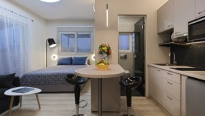 Een kleine keuken met een eethoek, een bed met blauw beddengoed, en een tafeltje met krukken.
