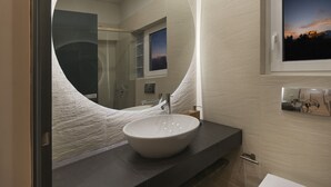Un baño moderno con un espejo redondo grande, un lavamanos blanco y una ventana con vista al atardecer.