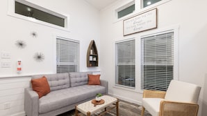 A living room with a grey sofa, a small wooden table with a plant, a white chair, and a wall-mounted shelf with books and a plant. - Oceans RV Resort (Holly Ridge)