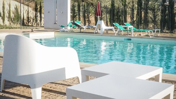 Zona de piscina con sillas de descanso blancas y una mesa, rodeada de árboles y un edificio al fondo.