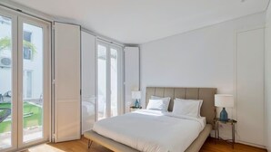 A modern bedroom with a large bed, a nightstand, a lamp, and a sliding glass door leading to a balcony. - Vita Luxus (Setúbal)