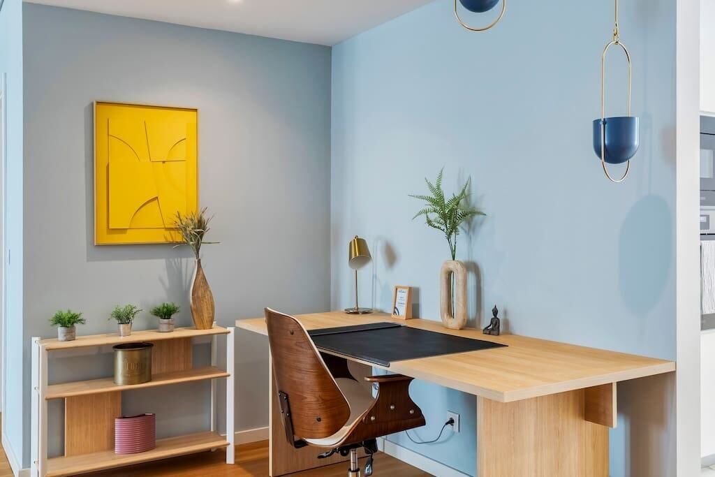 Ein moderner Arbeitsraum mit Schreibtisch aus Holz, einem Stuhl, einem Pflanzenregal, einem gelben abstrakten Gemälde und einer blauen Pendelleuchte.