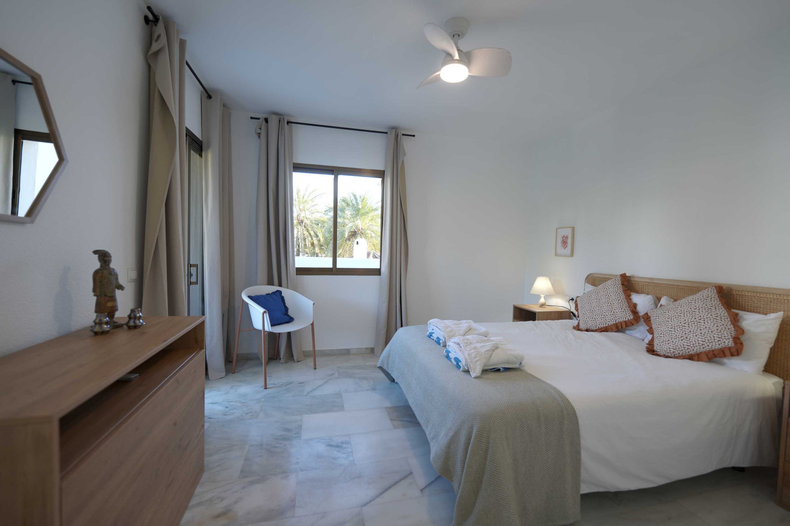 A hotel room with a bed, a chair, a desk, a ceiling fan, and a window with curtains.