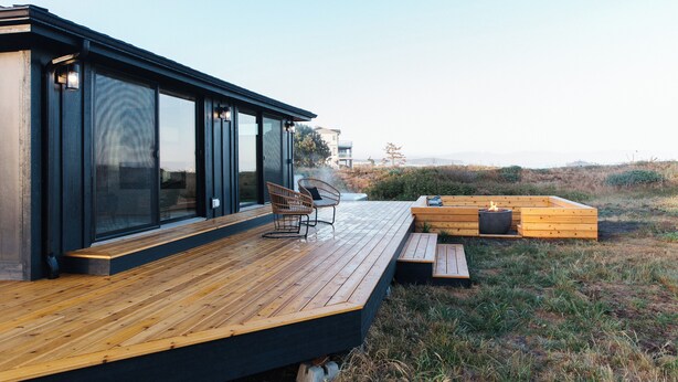 A modern outdoor deck with wooden flooring, a seating area with a fire pit, and large glass windows. - Lux Coastal Retreat & Hot Tub (Oak Harbor)