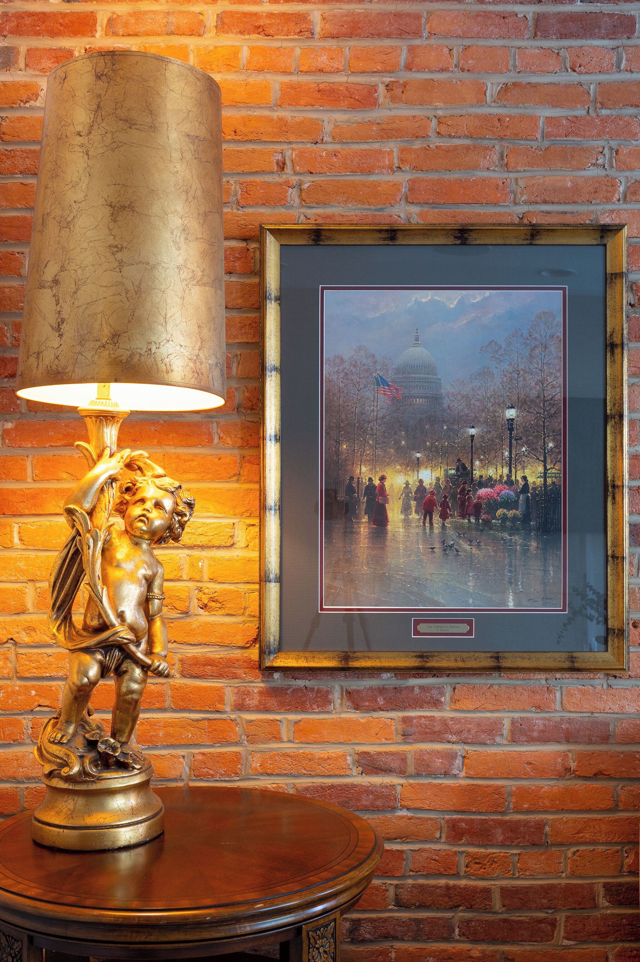 Eine Lampe mit zylindrischem Lampenschirm, ein gerahmtes Stadtbild und eine Backsteinwand.