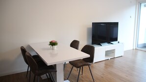 A modern living room with a white dining table, four brown chairs, a white entertainment unit with a flat-screen TV, and a potted plant. - Top Appartment in Hilden to rent, Lift, Fully furnished with equipped kitchen, (Hilden, Nordrhein-Westfalen, Deutschland)