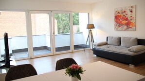A modern living room with a sofa, a dining table, and a large window with a view of a brick building and greenery. - Top Appartment in Hilden to rent, Lift, Fully furnished with equipped kitchen, (Hilden, Nordrhein-Westfalen, Deutschland)